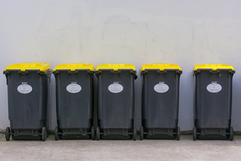 A row of five black plastic wheeled rubbish bins with yellow lids, lined up against a plain white wall on a paved outdoor surface. Each bin has a white label with a barcode and identification numbers