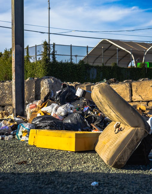 A large mound of black rubbish bags piled against a building wall featuring beige tiles, with some partially torn open revealing plastic bottles and paper waste inside. The bags are tightly knotted an