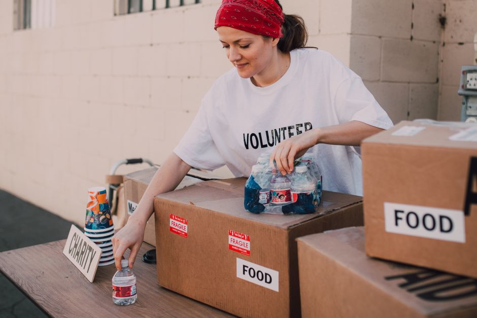 A woman wearing a white volunteer t-shirt and a red bandana is seen sorting and placing plastic bottles into a box labeled 'FOOD' on a wooden table outdoors. She is reaching for a small clear plastic