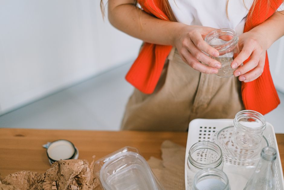 A person wearing a white shirt and an orange cardigan is holding a small glass jar with clear liquid, positioning their hands over a wooden table. On the table, there are multiple empty glass jars of 